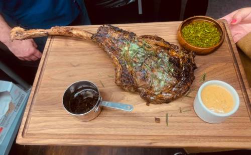 A large, grilled tomahawk steak on a wooden platter, accompanied by sauces and herbs.