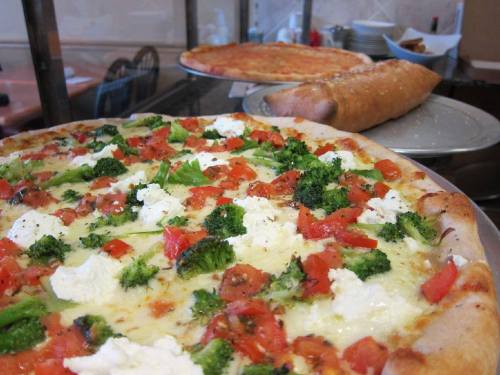 A close-up of a vegetable pizza topped with broccoli, tomatoes, and ricotta, with another pizza and breadsticks in the background.