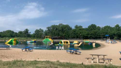 A sandy beach area with inflatable water attractions and picnic tables by a calm lake under a clear blue sky.