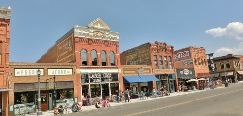 Historic brick buildings line a sunny street, featuring shops and outdoor seating, with people enjoying the day.