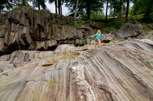A child playfully walks on smooth, layered rock formations surrounded by trees in a natural setting.