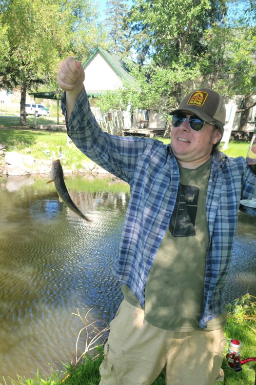 A person in a plaid shirt and cap holds up a fish by a pond, smiling in a sunny outdoor setting.