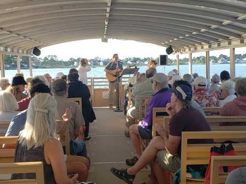 A musician performs on a boat, with an audience enjoying the music against a scenic waterfront backdrop.