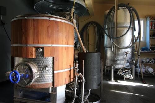 A wooden and metal brewing vessel in a workshop, with another metal tank and hoses in the background.