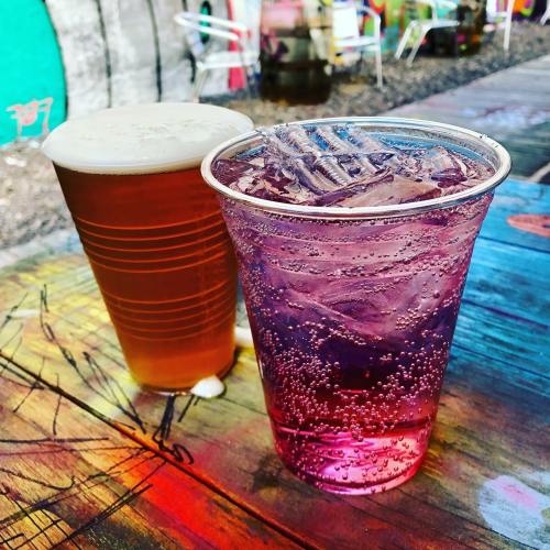 Two drinks on a colorful table: a frothy beer in a cup and a sparkling purple beverage with ice.