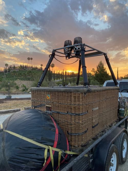 A hot air balloon basket and burner on a trailer, with a sunset and palm trees in the background.