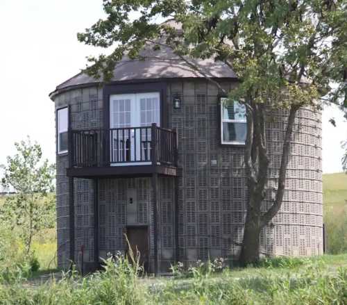 A unique round building with a balcony, surrounded by greenery and trees, resembling a grain silo.