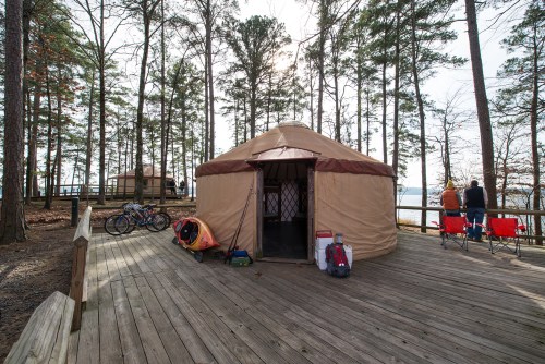 A yurt by a lake surrounded by trees, with bikes and kayaks nearby, and two people sitting on chairs.