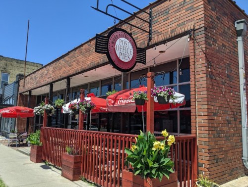 Exterior of a brick restaurant named "Post Office Pies," featuring outdoor seating with umbrellas and colorful flower planters.