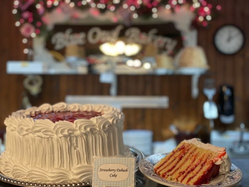 A beautifully decorated strawberry shortcake with a slice cut out, displayed in a bakery setting.