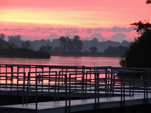 A serene river at sunrise, with vibrant pink and orange skies reflecting on the water and a wooden dock in the foreground.
