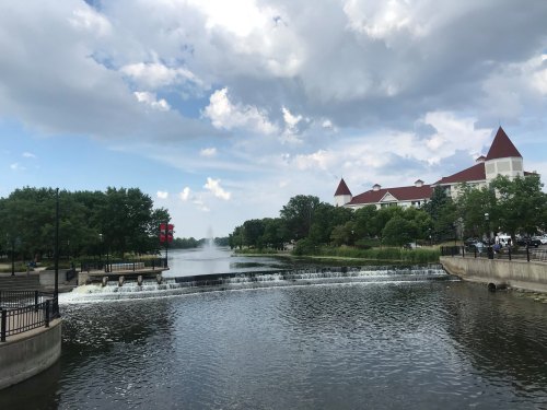 A serene river scene with a small waterfall, lush trees, and a building with a red roof under a cloudy sky.