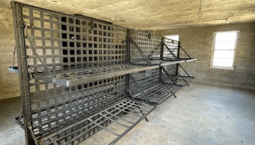 A dimly lit room with metal prison bunks against the walls and windows, featuring a concrete floor and textured walls.