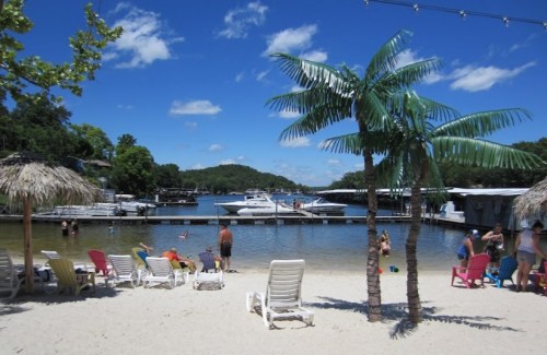 A sunny beach scene with palm trees, colorful lounge chairs, and boats docked on a calm lake.