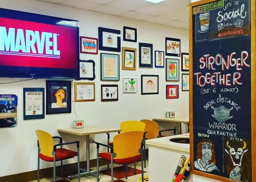 A cozy café with Marvel-themed art on the walls and a chalkboard promoting social distancing and community.