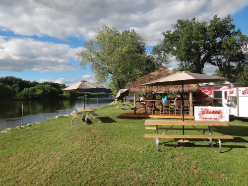 A riverside scene featuring a thatched-roof hut, picnic tables, and umbrellas under a partly cloudy sky.