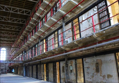 Interior of an abandoned prison, featuring peeling walls, barred windows, and a long corridor with multiple cells.