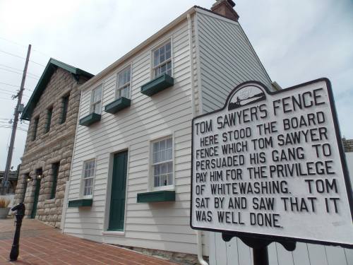 Historic sign in front of a white building, detailing Tom Sawyer's fence and his cleverness in persuading his friends.