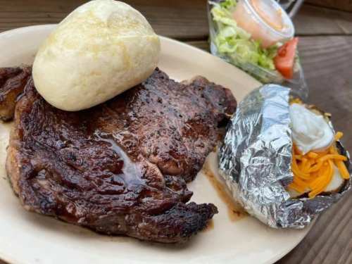 A plate featuring a grilled steak, a soft roll, a baked potato with cheese, and a side salad with tomato.
