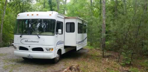 A white RV parked on a gravel path surrounded by lush green trees in a forested area.