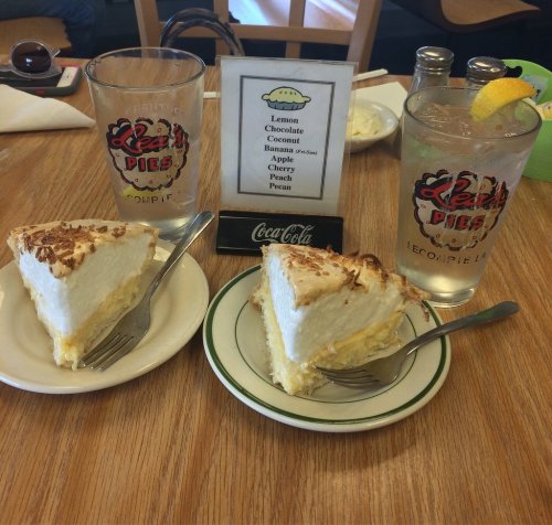Two slices of pie on plates, with drinks and a menu displaying pie flavors in a cozy diner setting.