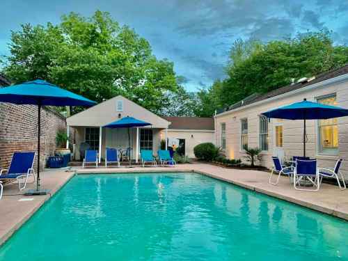 A serene pool area with blue umbrellas, lounge chairs, and greenery under a cloudy sky.