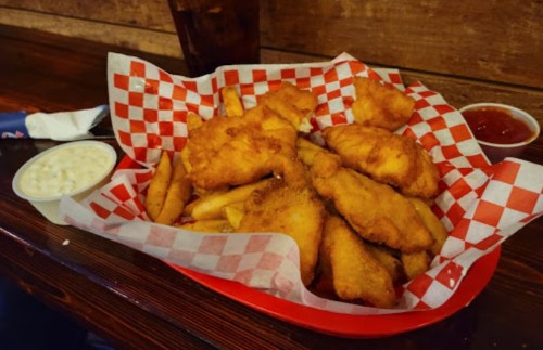 A basket of golden fried fish and fries, served with a side of coleslaw and ketchup on a checkered paper.