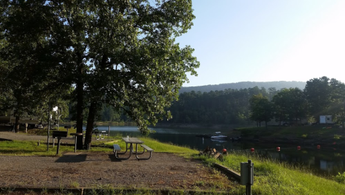 A serene lakeside view with picnic tables, trees, and a calm water surface reflecting the morning light.