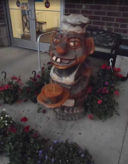 A wooden sculpture of a smiling chef holding a bowl, surrounded by colorful flowers near a building entrance.