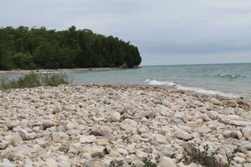 Rocky shoreline with gentle waves, surrounded by green trees and a cloudy sky.