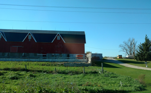 A red barn beside a grassy field, with a gravel path leading to a nearby building and trees in the background.