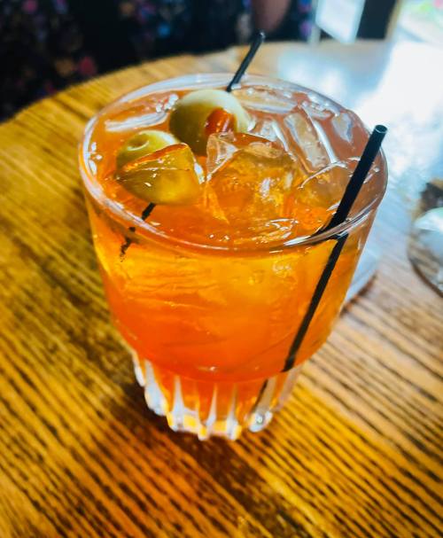 A vibrant orange cocktail with ice and two olives, served in a short glass on a wooden table.
