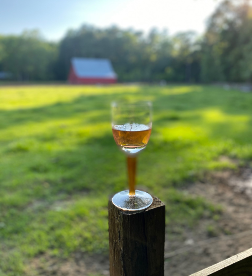 A glass of amber liquid sits on a wooden post, with a blurred green field and a red barn in the background.