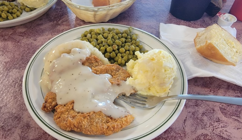 A plate of fried chicken with gravy, mashed potatoes, green peas, and a slice of bread on the side.