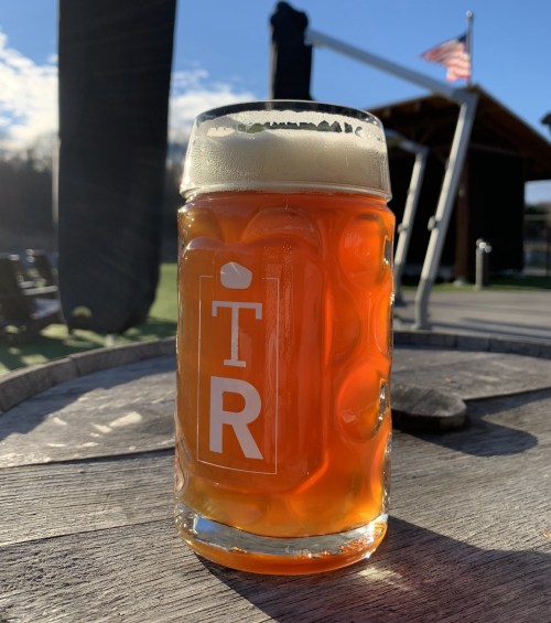 A glass of amber beer with a frothy head sits on a wooden table, with a flag and building in the background.