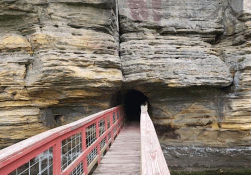 A wooden walkway leads into a dark tunnel carved into a rocky cliff, surrounded by textured stone walls.