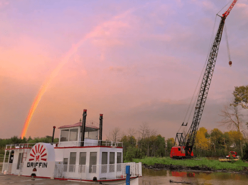 A barge named "Griffin" on water with a crane nearby and a rainbow in the sky during sunset.