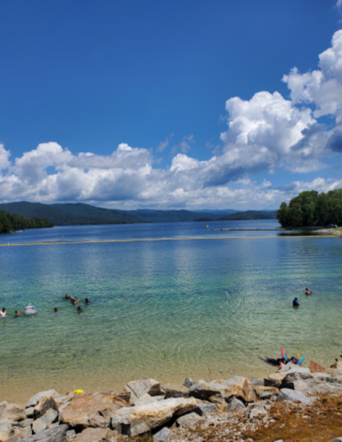 A serene lake with clear blue water, surrounded by green trees and fluffy clouds in a bright sky. People enjoy the water.
