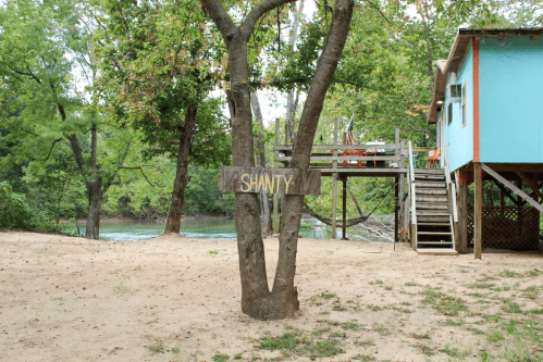 A wooden sign reading "SHANTY" hangs on a tree near a blue building by a river, surrounded by greenery.