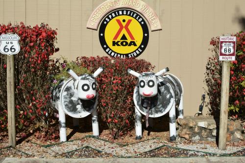 Two cow sculptures with black and white spots stand in front of a KOA sign, surrounded by colorful foliage.
