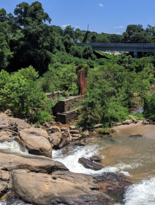 A scenic view of a river with rocky banks, lush greenery, and remnants of old stone structures under a clear blue sky.