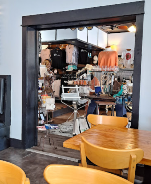 A view of a boutique through an open doorway, showcasing clothing and accessories, with wooden chairs in the foreground.