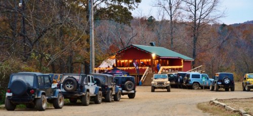 A rustic building with festive lights surrounded by parked off-road vehicles on a dirt road in a wooded area.