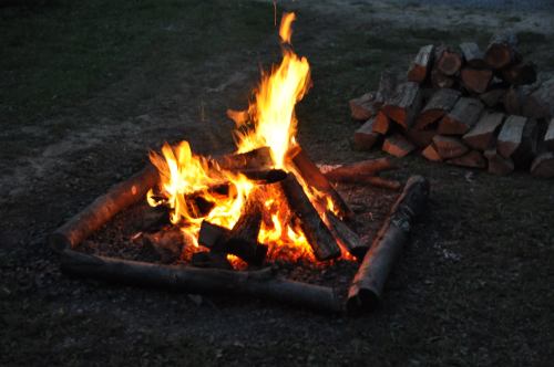 A campfire burns brightly in a circular arrangement of logs, with a stack of firewood nearby on the ground.