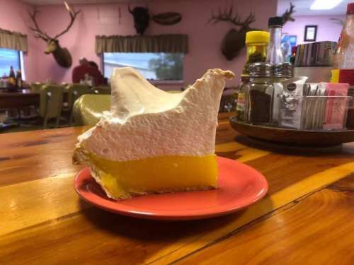 A slice of pie with a yellow filling and white meringue on a pink plate, set on a wooden table in a diner.