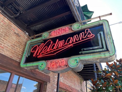 Neon sign for Weldmann's, established in 1870, mounted on a brick wall with a clear blue sky above.