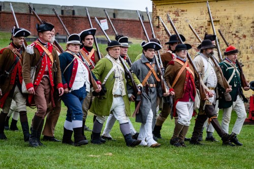 A group of historical reenactors in 18th-century military uniforms marching together on a grassy field.