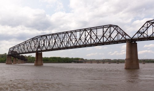A large metal bridge spans a river, with cloudy skies and green trees in the background.
