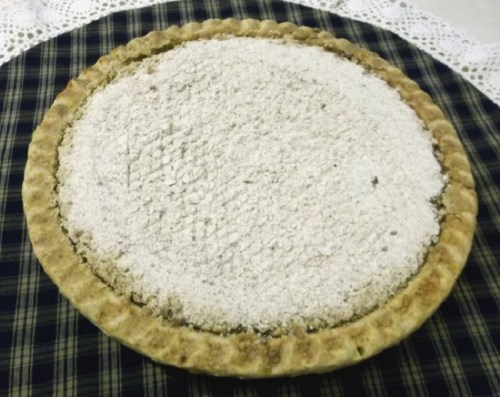 A round pie with a crumbly, powdered sugar topping on a lace and plaid tablecloth.