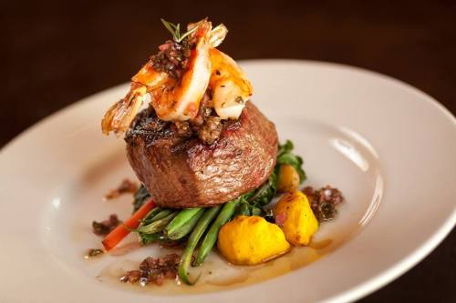 A gourmet dish featuring a grilled steak topped with shrimp, served on a bed of green beans and yellow squash.